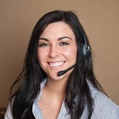 A woman with long dark hair is smiling and wearing a headset. She is dressed in a striped shirt and is positioned against a plain beige background